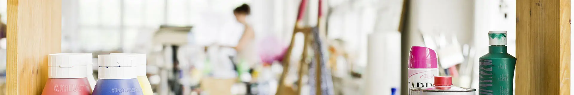 Das Bild zeigt ein Regal mit verschiedenen Kunstmaterialien, darunter Farbtuben und Sprühdosen. Im Hintergrund ist ein Künstleratelier zu erkennen, mit verschwommenen Figuren und Gegenständen wie einer Staffelei und weiteren Malutensilien. Auf den Flaschen sind Texte zu lesen, darunter „C.KREUL“ auf einer roten Flasche und „KÜNSTLERARBEIT“ auf einer blauen Flasche.