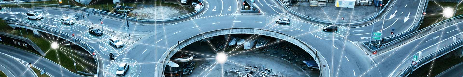 Das Bild zeigt eine Luftaufnahme eines komplexen Straßenkreuzes mit mehreren Ebenen und Fahrspuren. Auf den Straßen sind mehrere Autos und ein Motorrad unterwegs. Über das Bild sind digitale Grafiken gelegt, die Kommunikationssignale oder Vernetzung zwischen den Fahrzeugen darstellen – ein Hinweis auf ein intelligentes Verkehrssystem oder autonome Fahrzeugtechnologie.  Die Straßen sind mit üblichen Verkehrsschildern und Fahrbahnmarkierungen versehen. Im zentralen Bereich unter einer der erhöhten Fahrbahnen ist zudem etwas Baumaschinen-Equipment zu sehen.