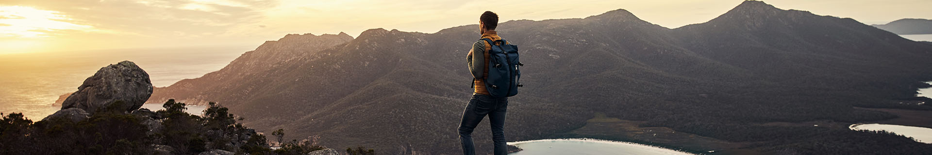 Das Bild zeigt eine Person, die auf einem felsigen Vorsprung steht und eine weite Landschaft aus Bergen und Tälern überblickt. Die Person trägt einen Rucksack und Outdoor-Kleidung, was auf eine Wanderung oder Erkundungstour hinweist. Die Szene spielt sich bei Sonnenaufgang oder Sonnenuntergang ab – der Himmel leuchtet in warmen Orangetönen und Gelbtönen.  Im Hintergrund erstrecken sich große Gebirgszüge, und in den Tälern ist etwas Vegetation zu erkennen. In der unteren rechten Ecke des Bildes ist teilweise ein Gewässer sichtbar.