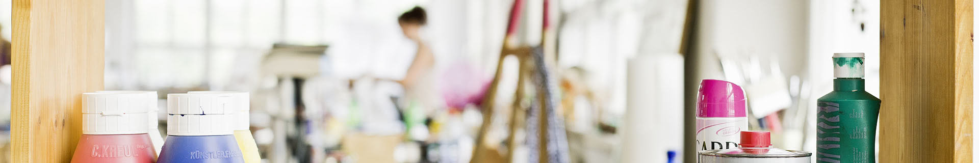 Das Bild zeigt ein Regal mit verschiedenen Kunstmaterialien, darunter Farbtuben und Sprühdosen. Im Hintergrund ist ein Künstleratelier zu erkennen, mit verschwommenen Figuren und Gegenständen wie einer Staffelei und weiteren Malutensilien. Auf den Flaschen sind Texte zu lesen, darunter „C.KREUL“ auf einer roten Flasche und „KÜNSTLERARBEIT“ auf einer blauen Flasche.