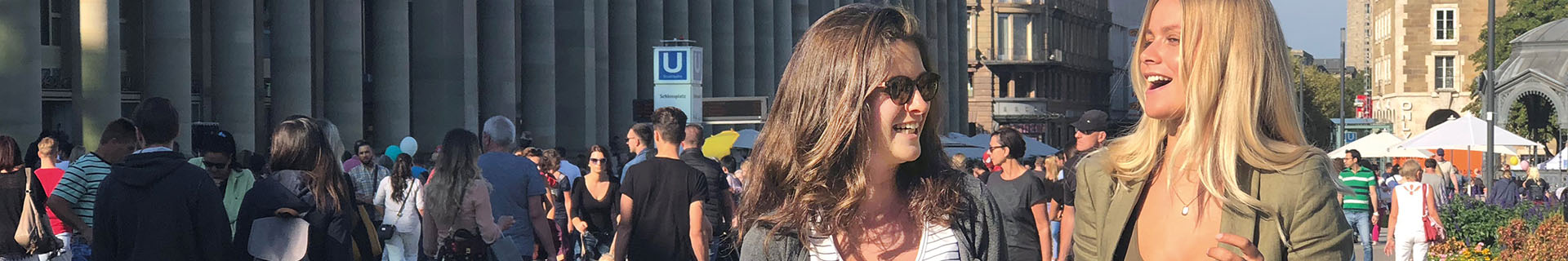 Das Bild zeigt eine belebte Szene im Freien mit vielen Menschen, die herumlaufen oder stehen. Im Hintergrund sind große Säulen zu sehen, vermutlich Teil eines bedeutenden Gebäudes oder einer öffentlichen Struktur. Ein Schild mit dem Buchstaben „U“ weist auf einen U-Bahn-Eingang hin. 